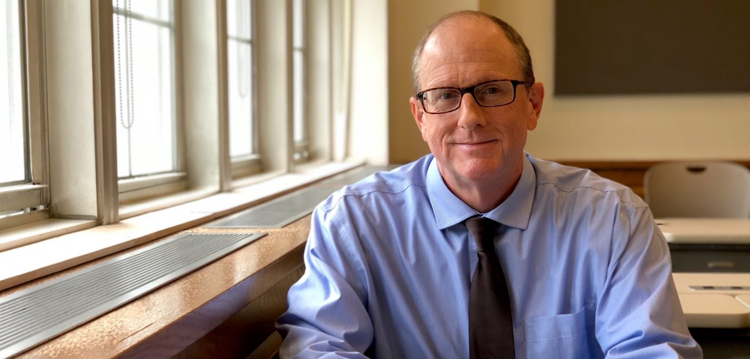 A portrait of C. Colt Anderson, sitting in a classroom and wearing a periwinkle blue shirt and tie.