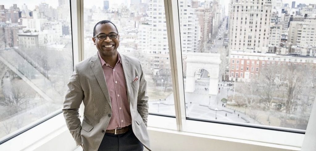 A man wearing glasses and a blazer smiles in front of a window.