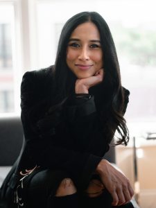 A woman wearing all black smiles at the camera with her chin propped under her hand.