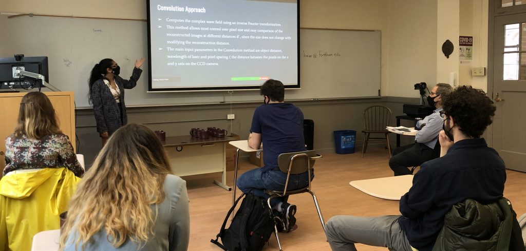 A woman gestures towards a presentation screen in a classroom full of people, spaced six feet apart.