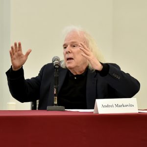 A man with white hair and a black jacket throws his arms up.