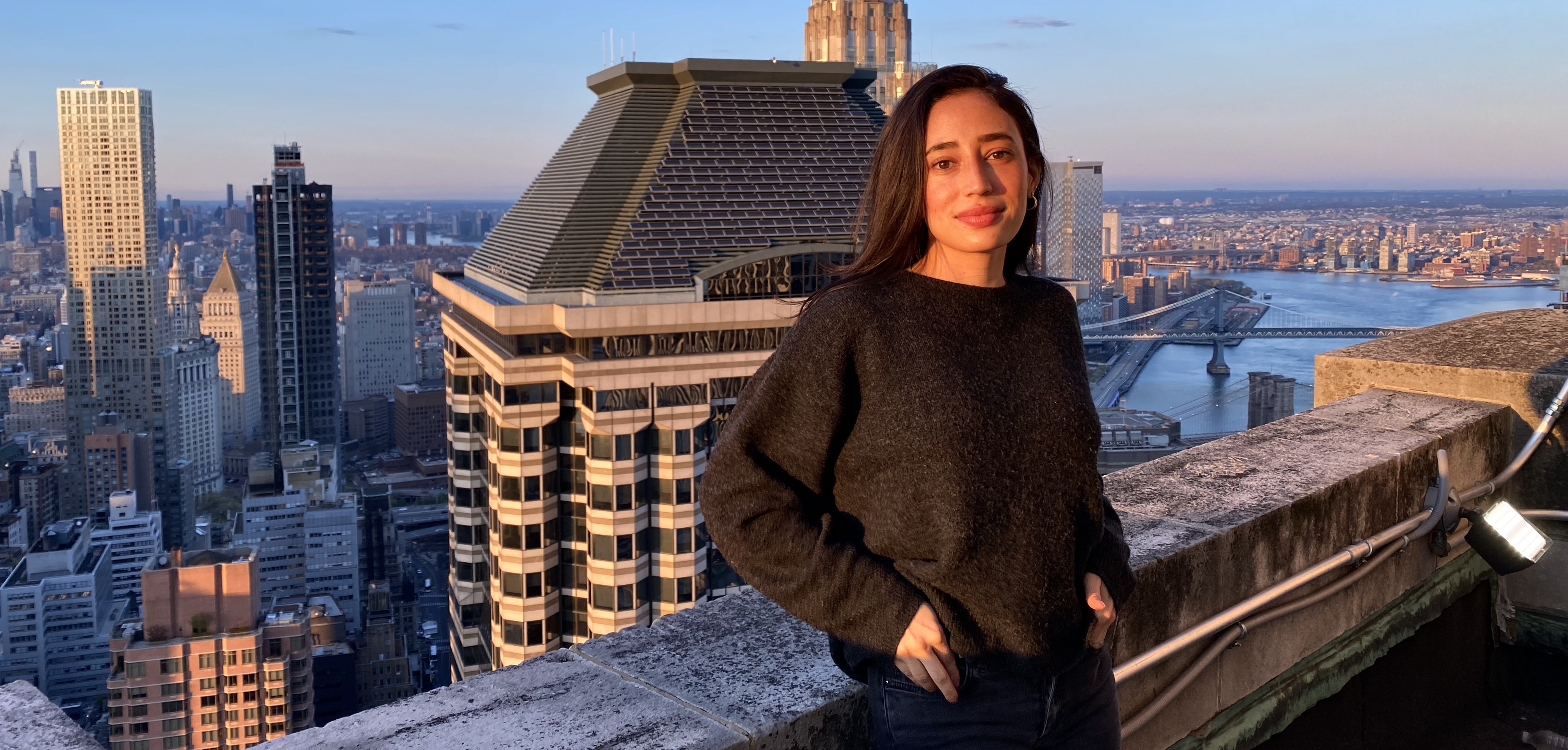 A woman wearing a black sweater stands in front of the Manhattan city skyline.