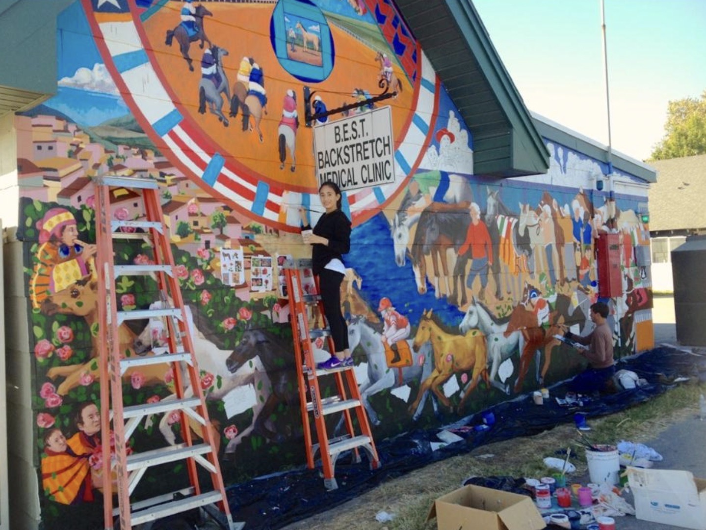 A woman stands on a ladder in front of a mural and smiles.