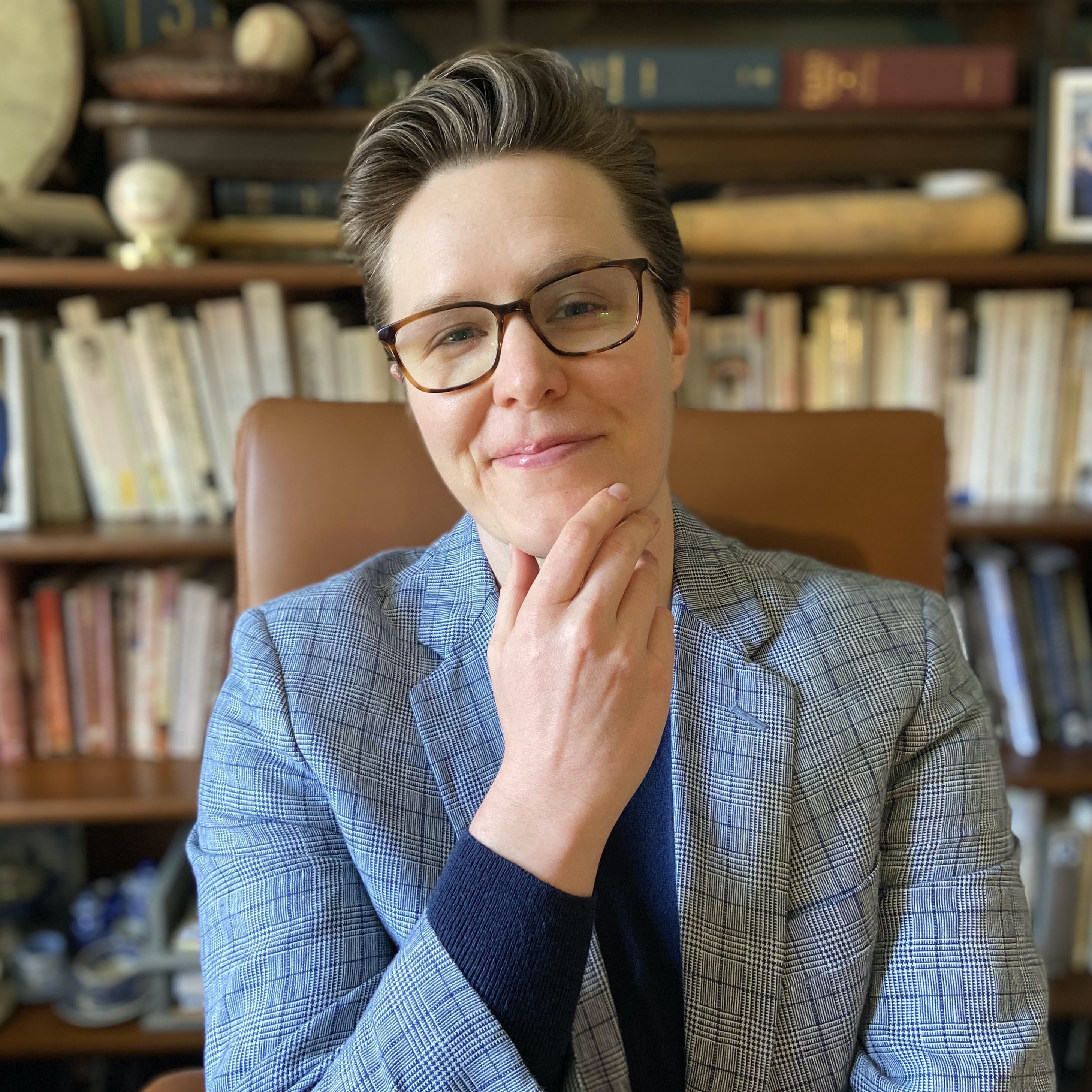 A woman wearing a blazer holds her chin in her hand in front of a bookshelf and smiles.