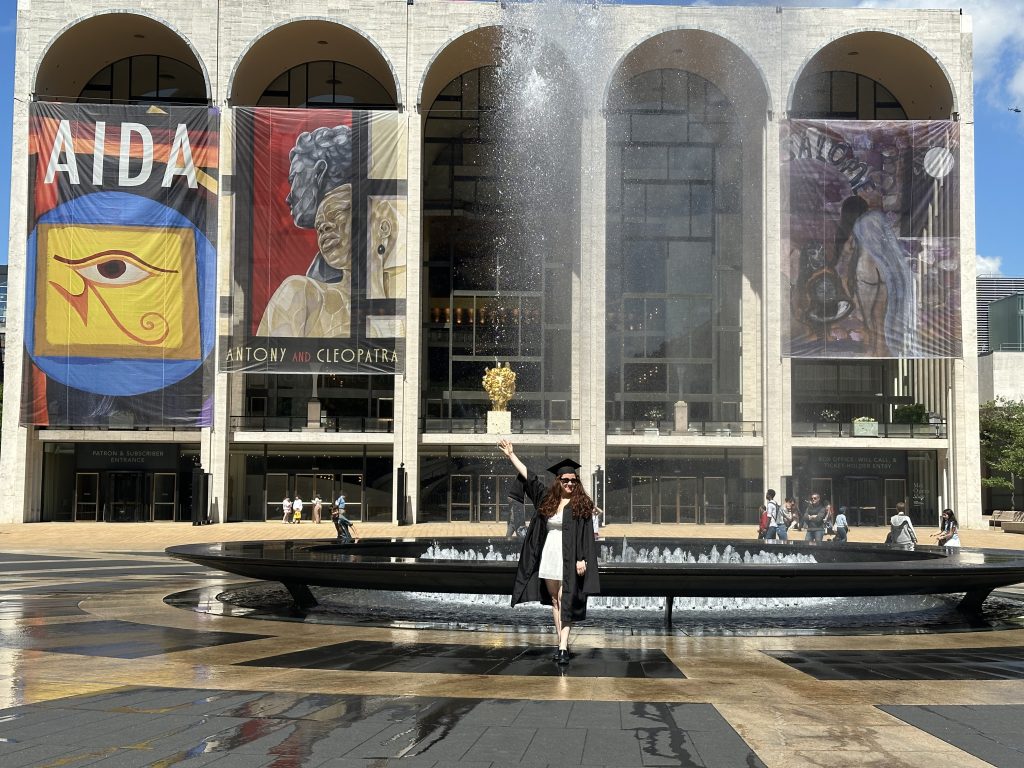 Woman with hand up in cap and gown in front of the fountain in the Lincoln Center for Performing Arts