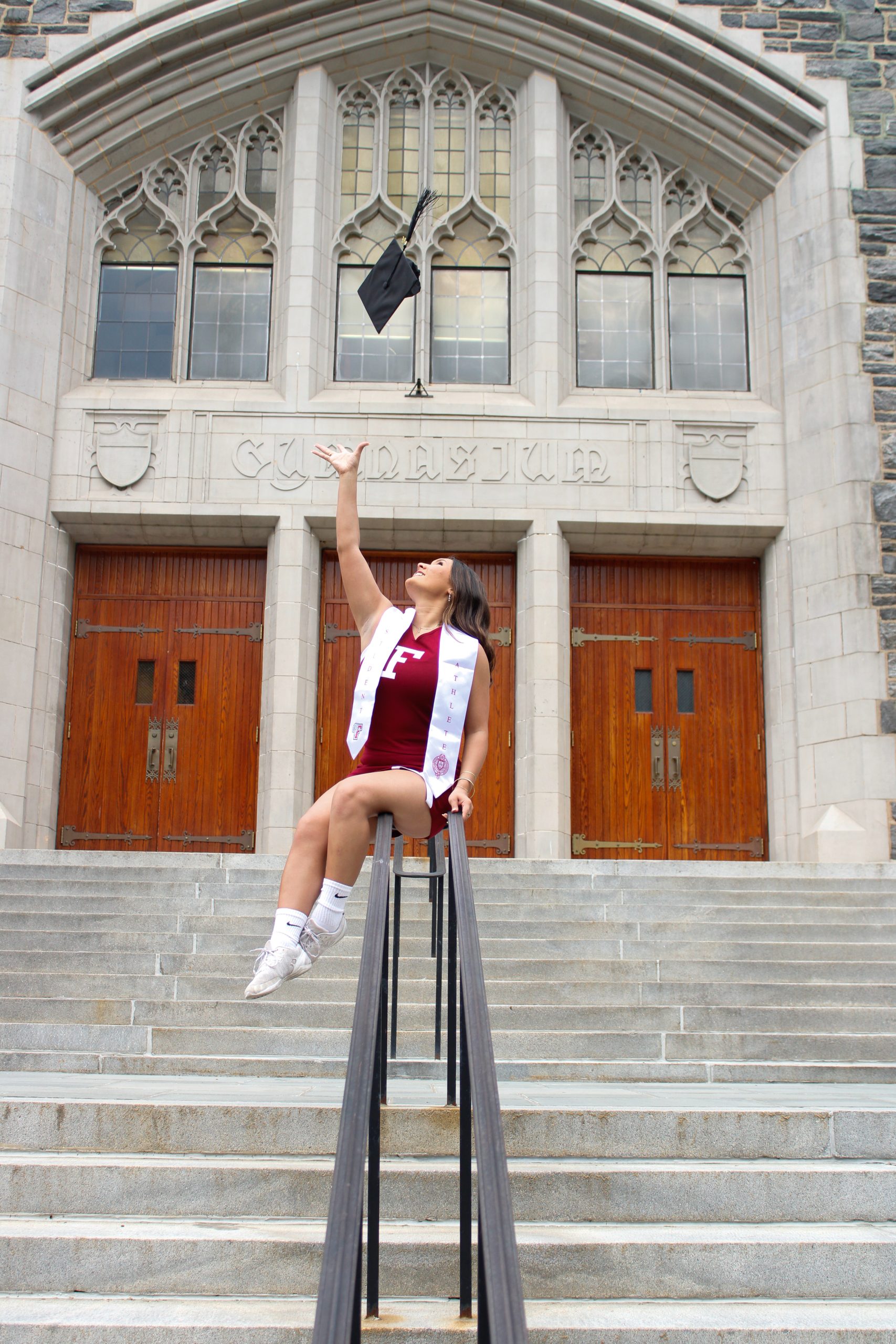 Woman throwing cap in air on gym steps (outside) she is siting on the hand rails.