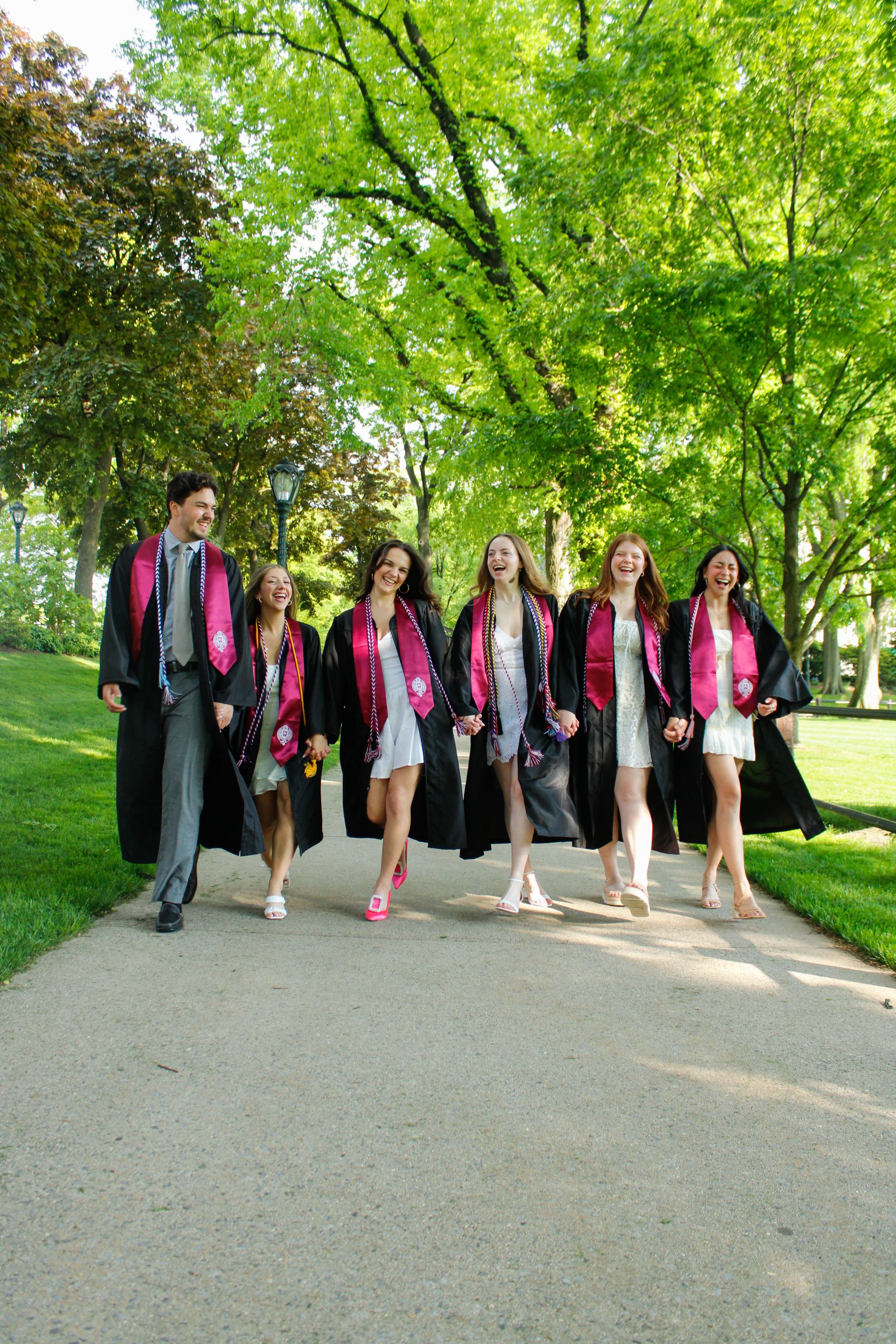 One man (left) 5 women (right) walking on sidewalk in graduation gowns