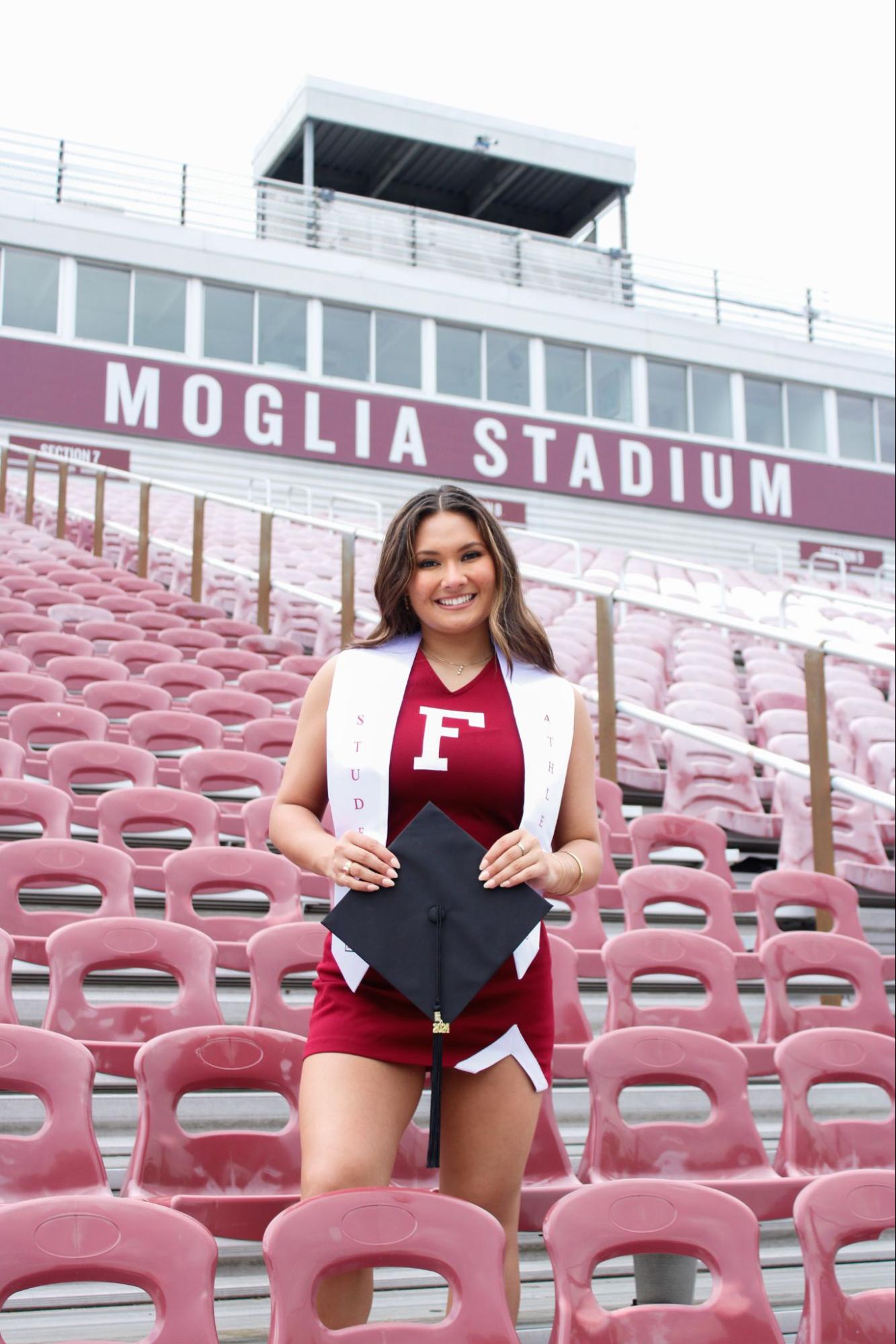 Woman standing alone in bleachers holding cap. Moglia Stadium sign in the background