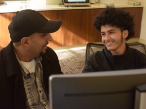 A son smiles at his father behind a computer screen.