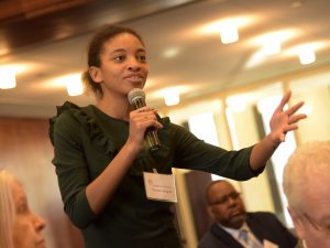 Victoria Draper extends her arm, speaking to the panelists with a microphone.