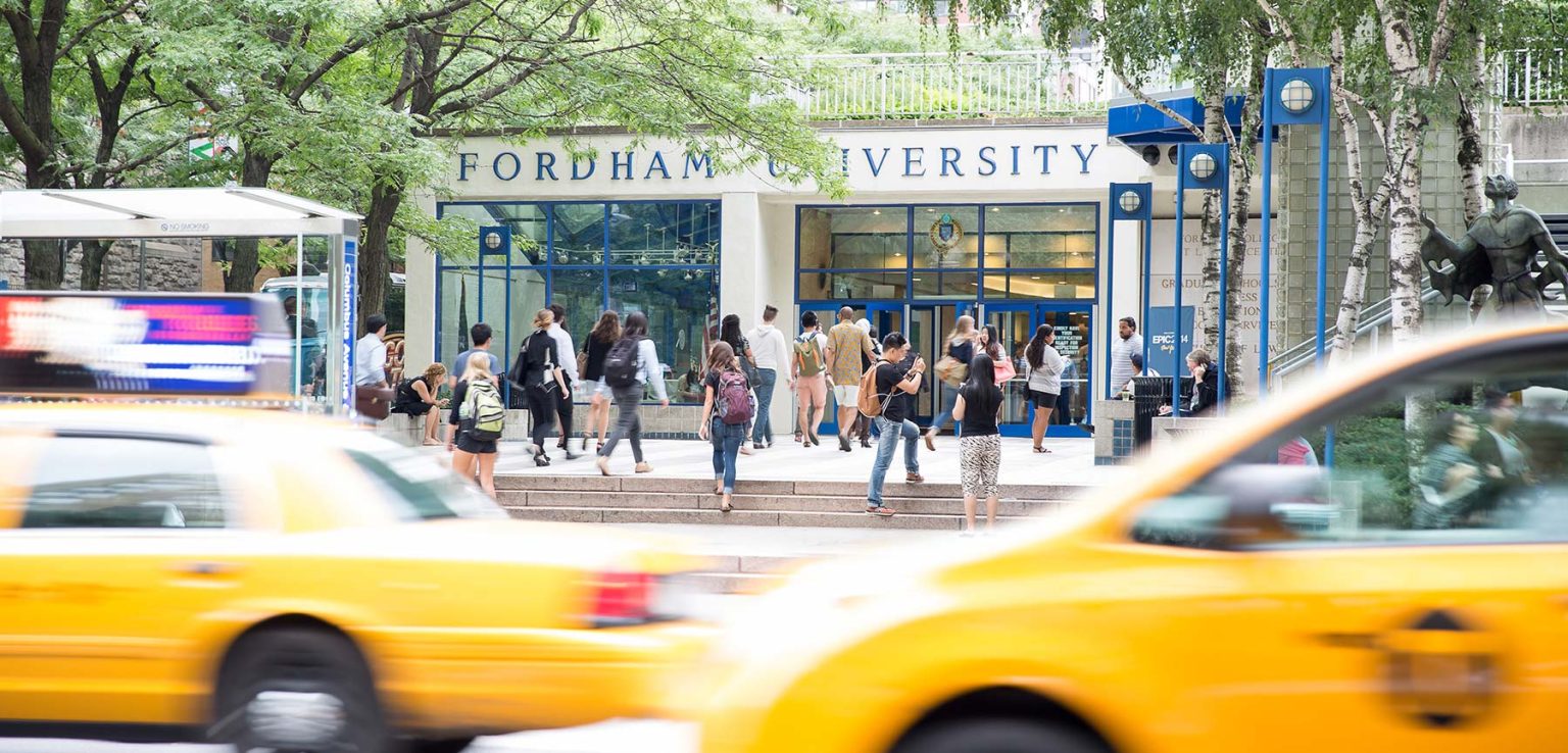 People walk in front of the entrance to Fordham's Lowenstein Center, as two yellow cabs pass in the foreground.