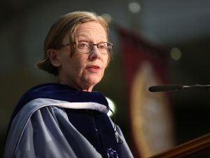 A woman wearing light blue graduation robes speaks before a microphone