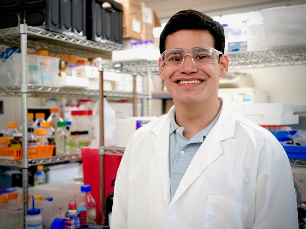 A young man wearing a white lab coat and goggles smiles at the camera