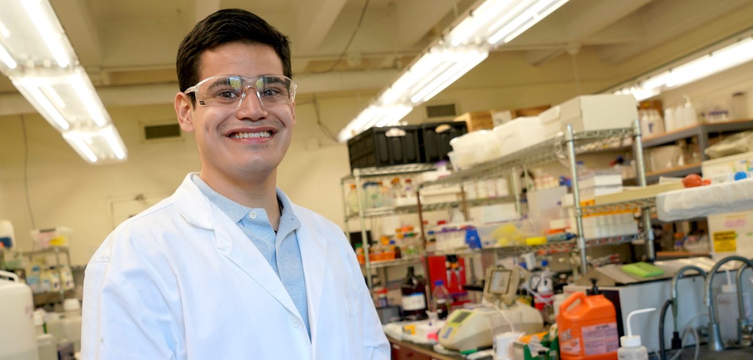 A young man wearing a white lab coat and goggles smiles at the camera