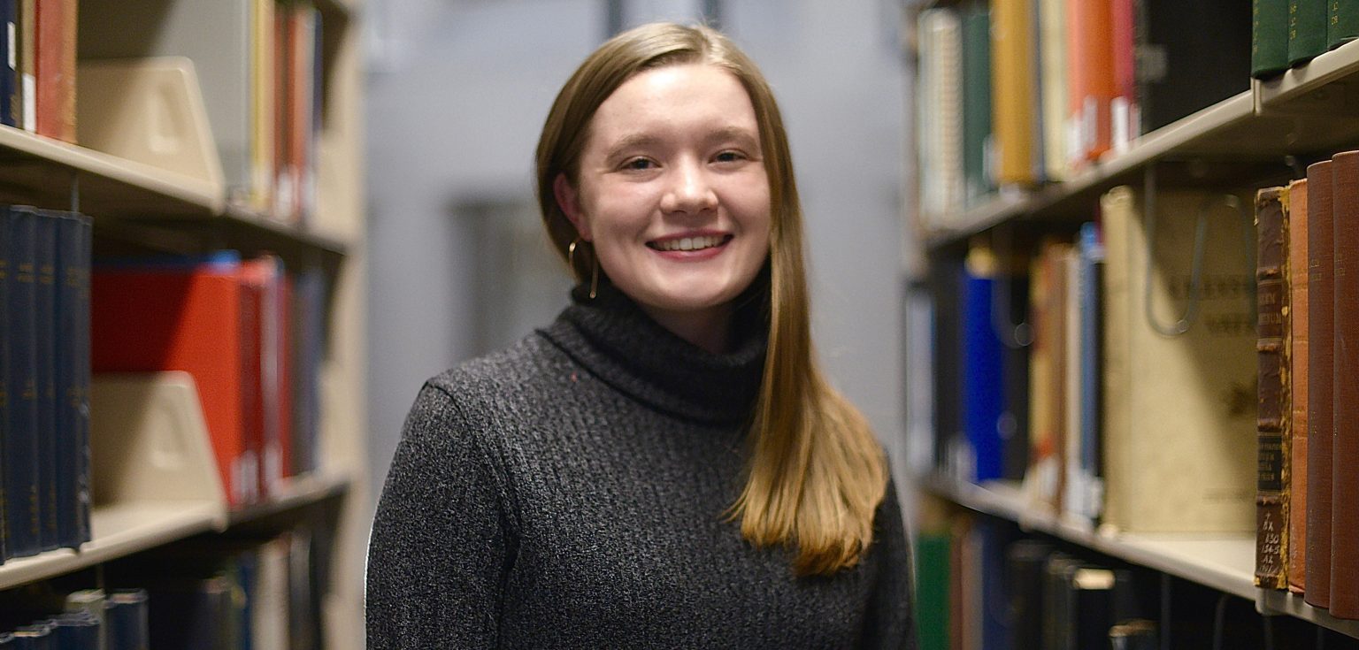 A blonde girls smiles in between two bookshelves