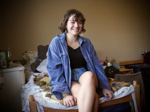 A girl wearing a denim jacket sits on a bed and smiles.