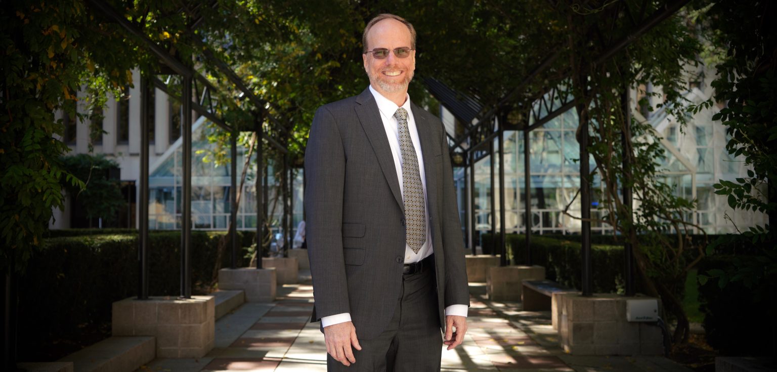 A man wearing glasses and a suit smiles in front of a grassy pathway.