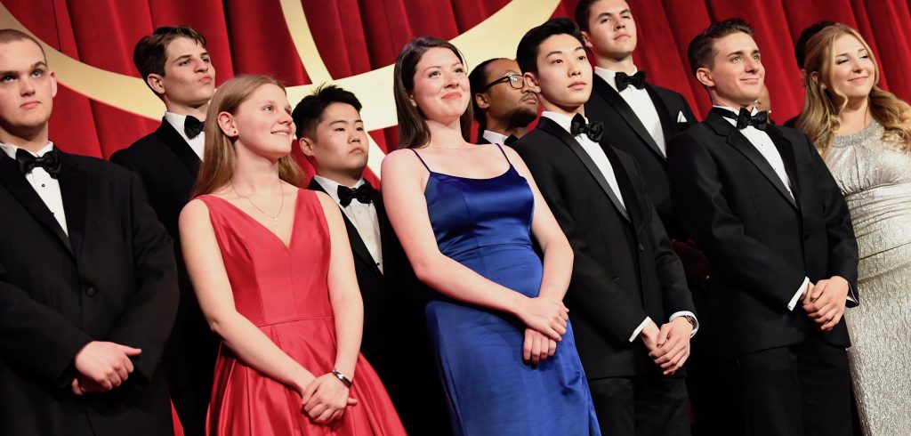 A group of young men and women dressed in gowns and suits stand together.