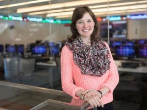 A young woman wearing a pink sweater smiles in front of computer terminals.