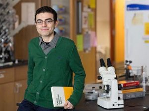 A young man wearing a green cardigan and glasses smiles in a science classroom.