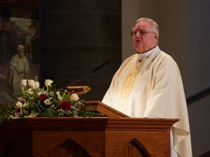 A man wearing white priestly garb stands at a podium.