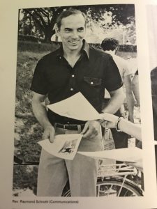 A black and white photo of a man standing, smiling, and holding a few papers