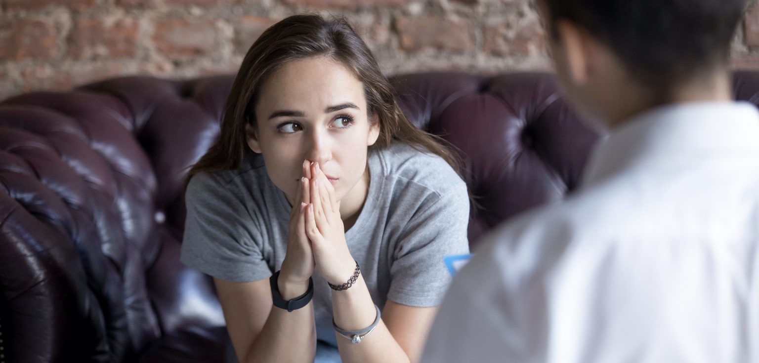 A young woman sits forward with her hands pressed together.