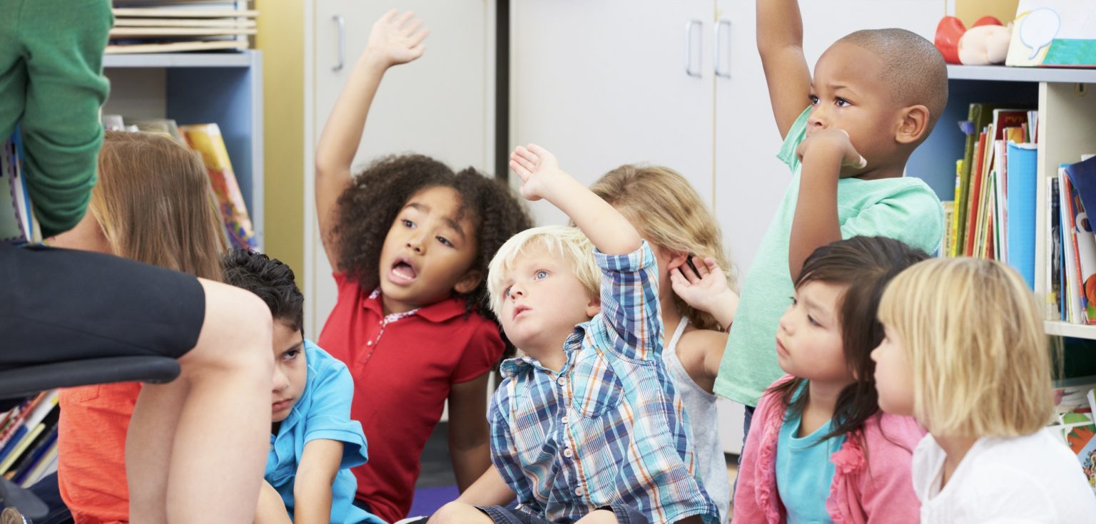 Children sit on a classroom carpet and excitedly raise their hands.