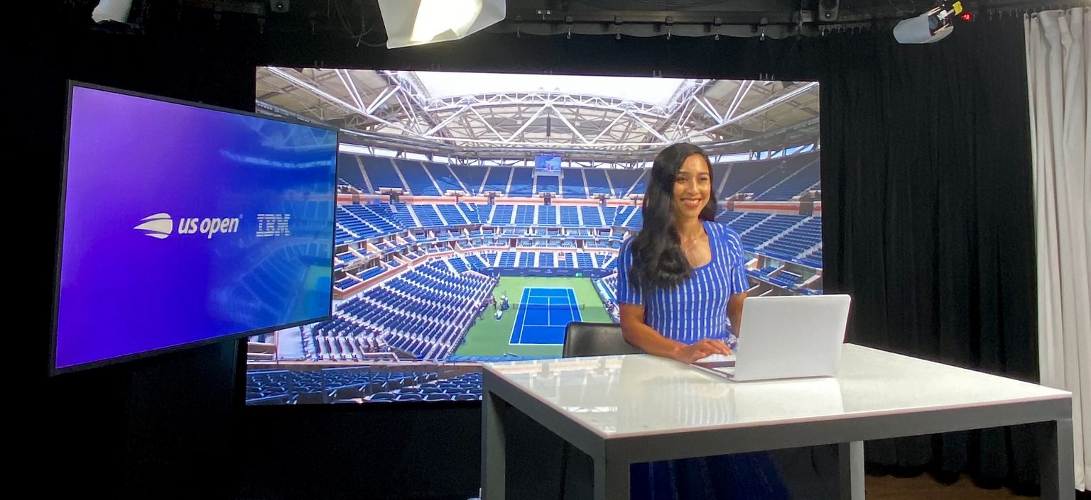 A woman wearing a blue dress smiles in front of a screen picturing an empty tennis court.