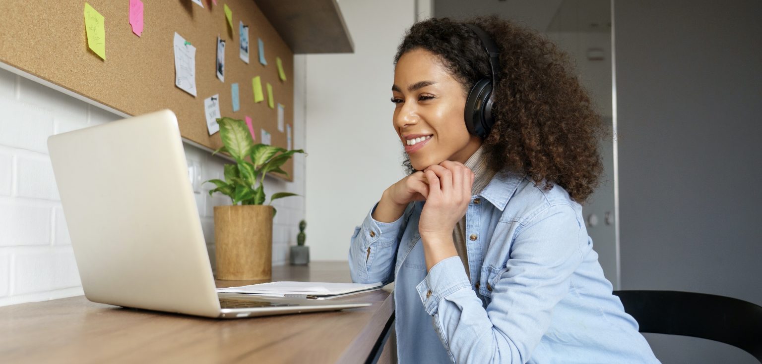 A woman with curly hair and a jean jacket is wearing headphones and is seated at a table in front of a laptop.