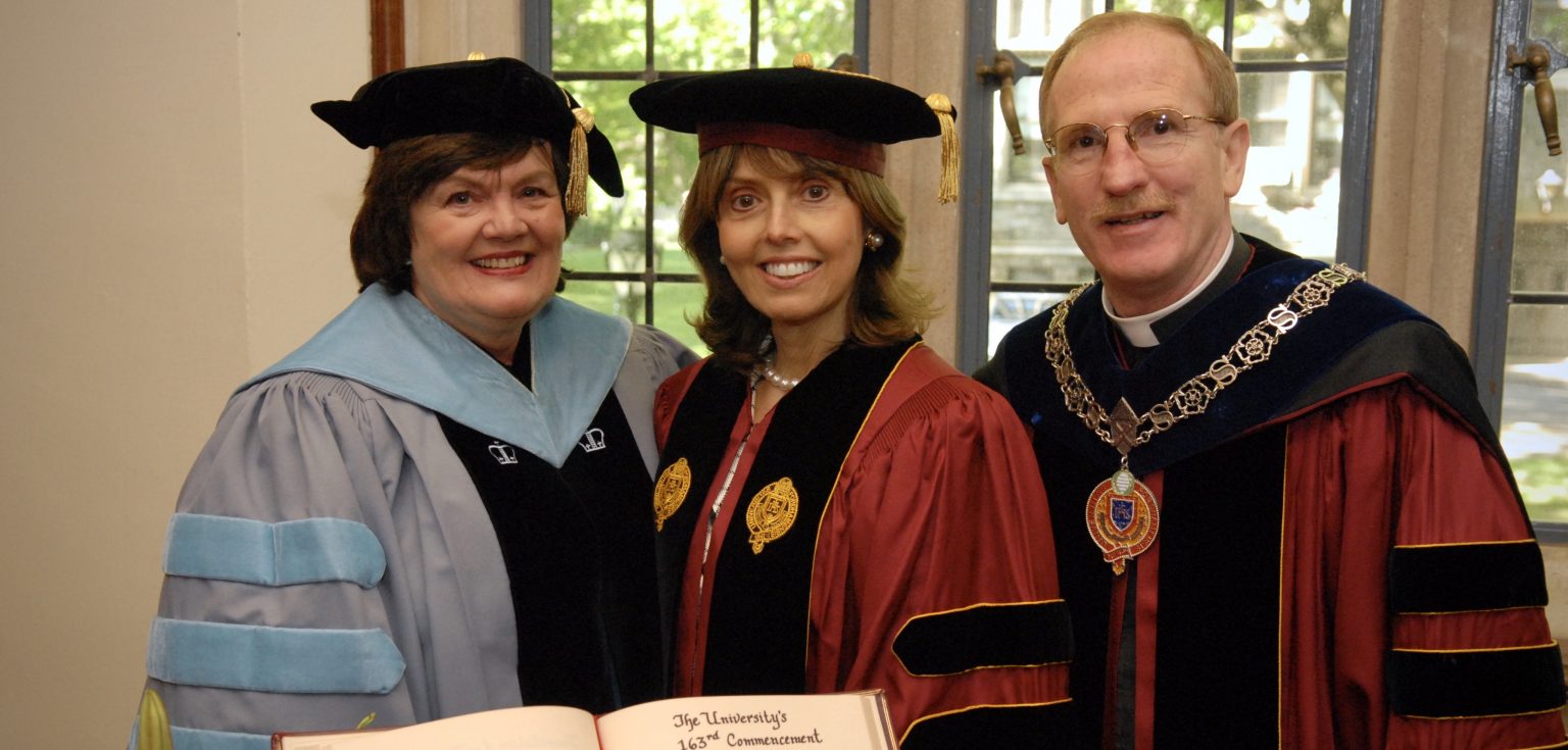 A woman wearing a blue graduation robe, a woman wearing a maroon graduation robe, and a man wearing a maroon graduation robe smile at the camera together.