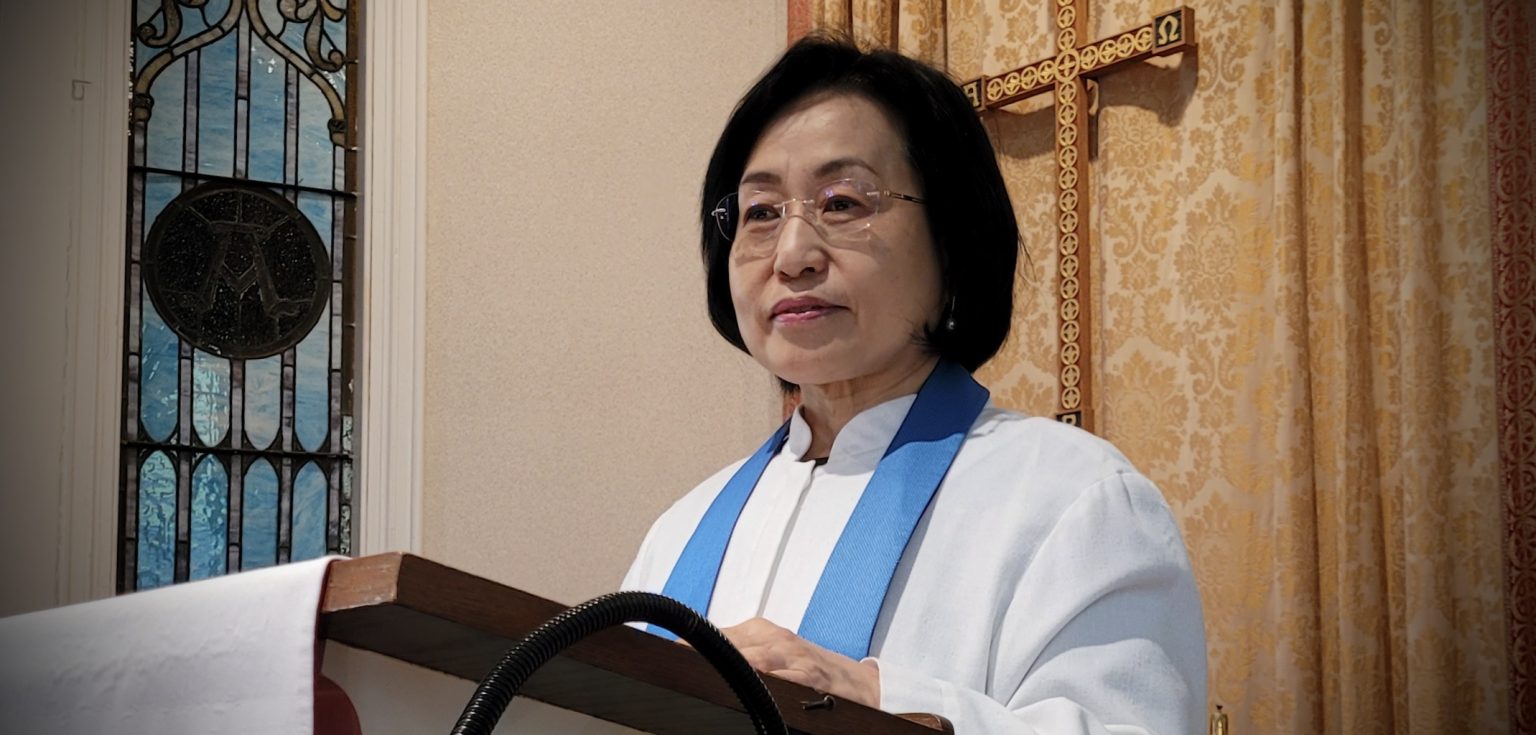 An Asian-American woman wearing glasses and a white pastor's robe stands in front of a pulpit.