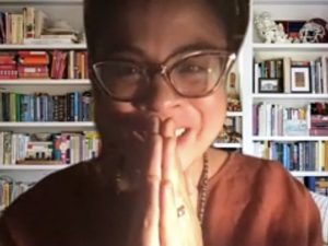 A woman wearing glasses clasps her hands in front of a background filled with books.