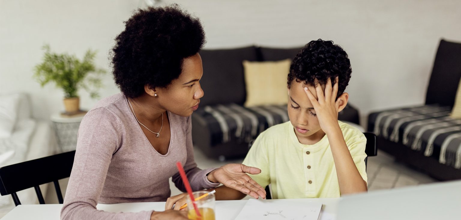 A woman with an afro and light pink shirt speaks to a boy with curly hair and a yellow T-shirt at a white desk.