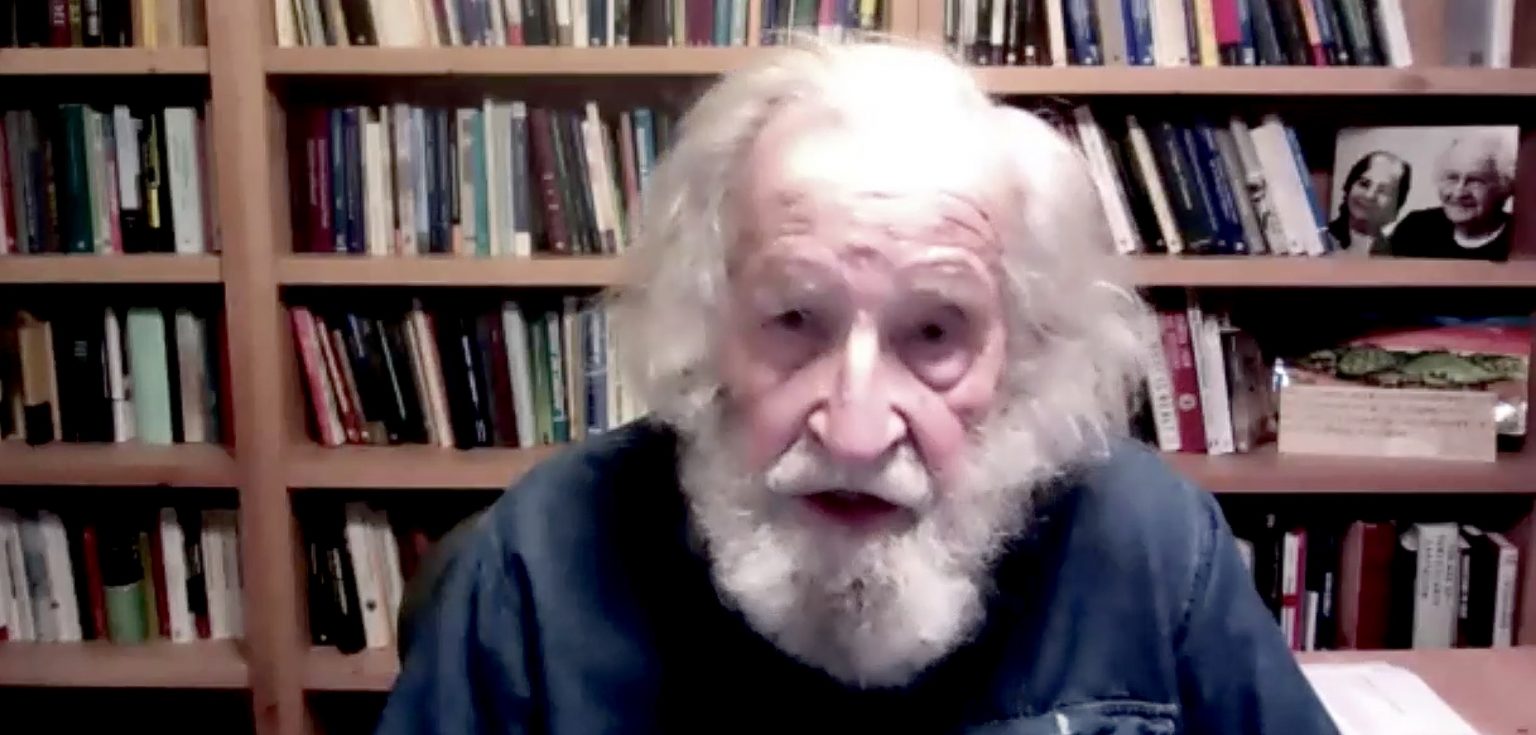 A man with white head and beard hair talks in front of a bookshelf