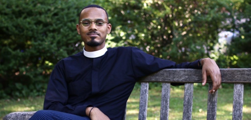 A man wearing glasses and black priest robes sits on an outdoor bench and smiles at the camera.