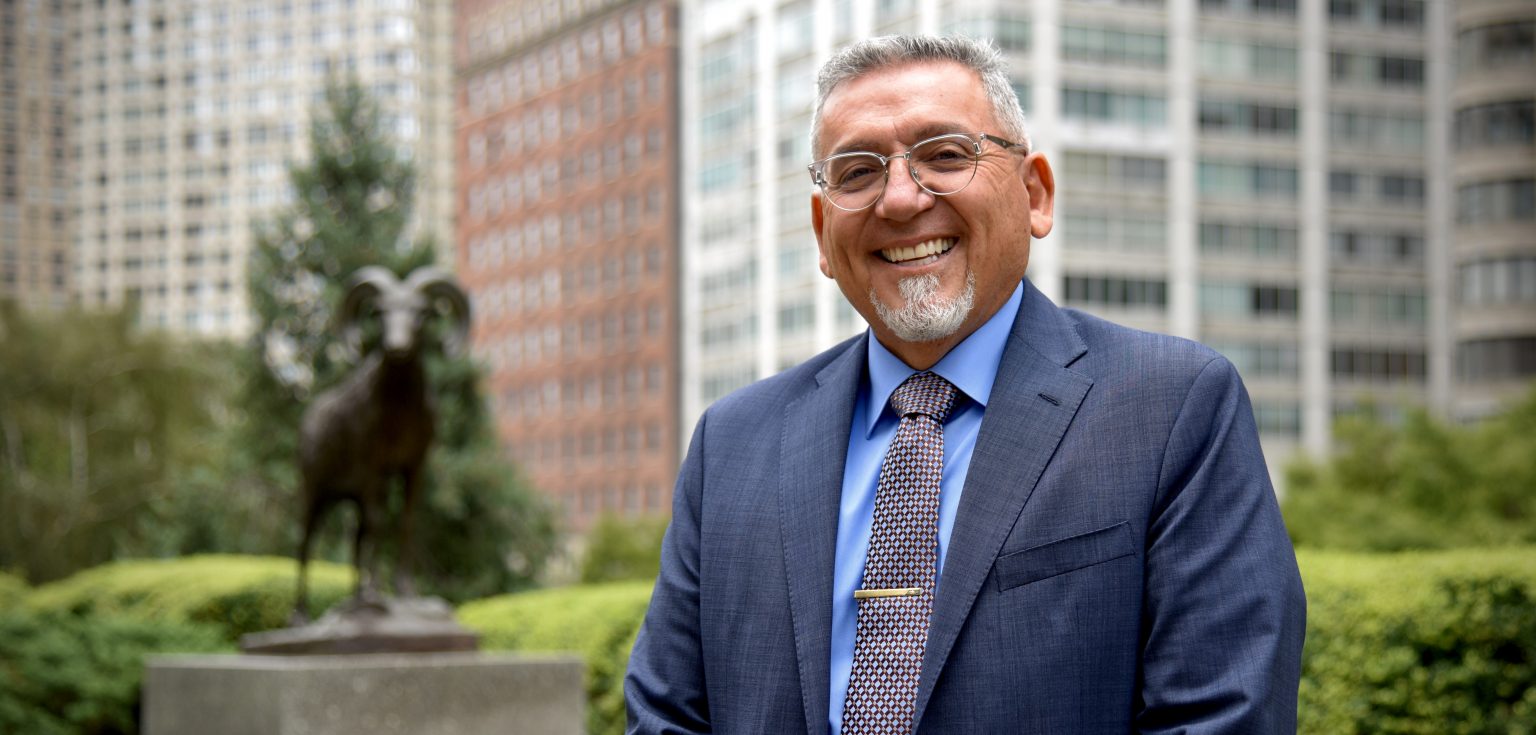 A man wearing glasses, a navy blue blazer, and a patterned tie smiles at the camera.