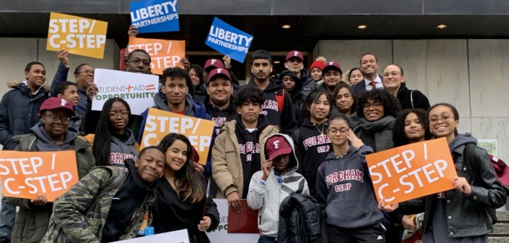 A group of young adults wearing winter coats and holding colorful signs smile for the camera.