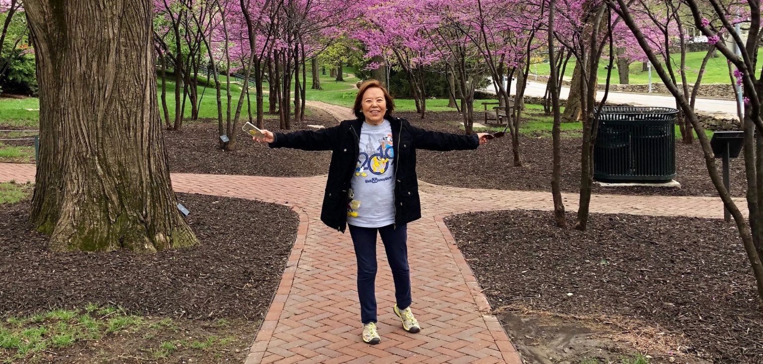 An elderly woman of Asian descent smiles and stands in front of a group of trees with pink blossoms while throwing out her arms.