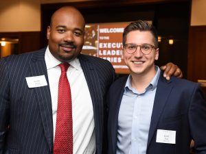 Two men wearing blazers smile next to each other.