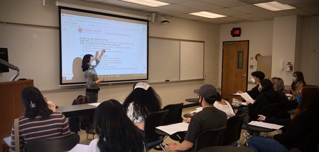 A woman wearing a mask gestures at a projector with Korean letters on it in front of a classroom full of students.