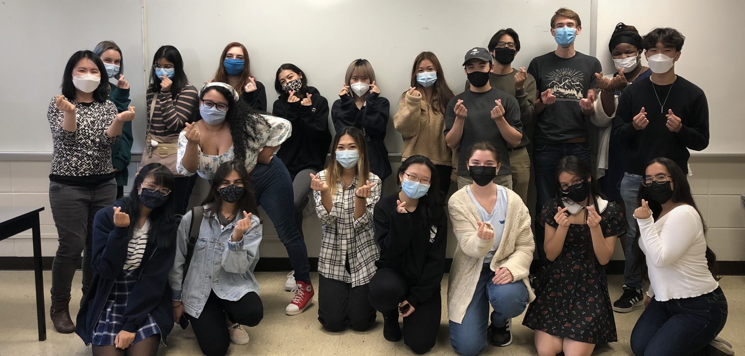 Twenty people wearing masks smile at the camera in front of a blank dry erase board and pose with their fingers in the shape of a heart.