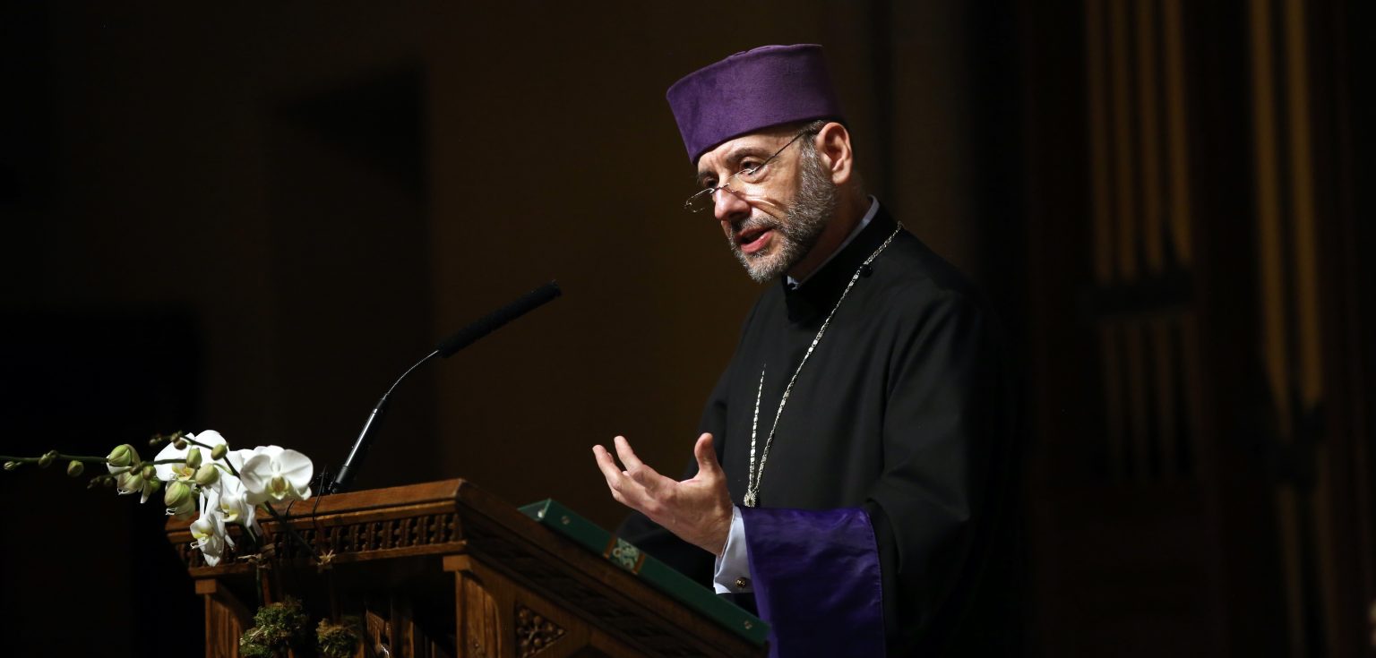 A man wearing a purple hat and black priestly robes holds his hand up at a podium.