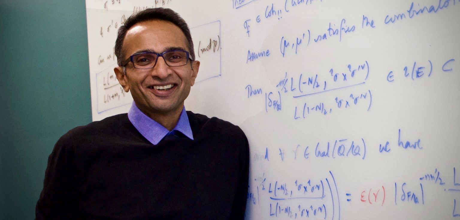 A man wearing glasses smiles in front of a board filled with math equations.