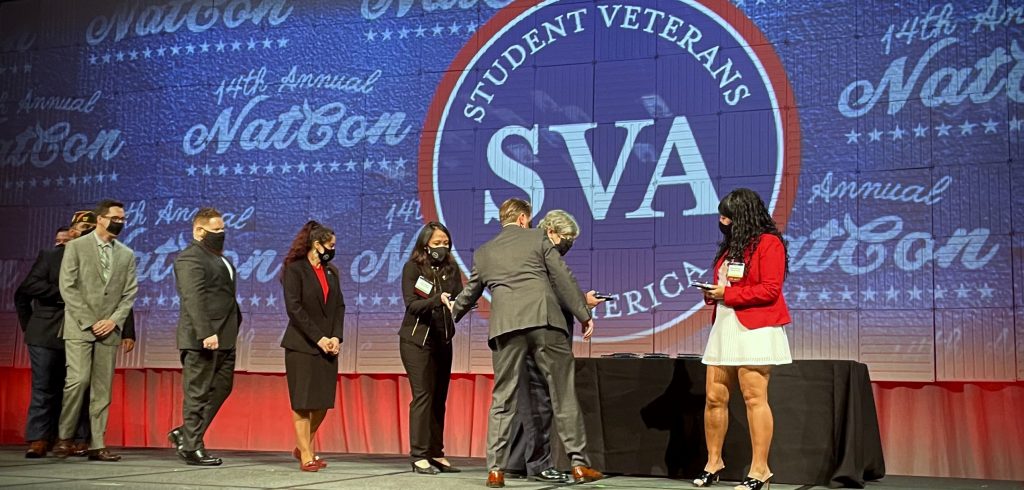 A line of people wearing masks get ready to shake hands with someone on a large stage.