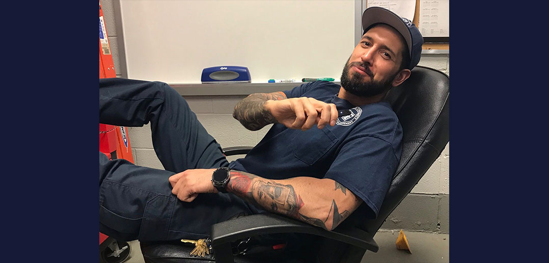 A man wearing a dark blue T-shirt with tattoos and a baseball cap slouches in a chair and smiles.