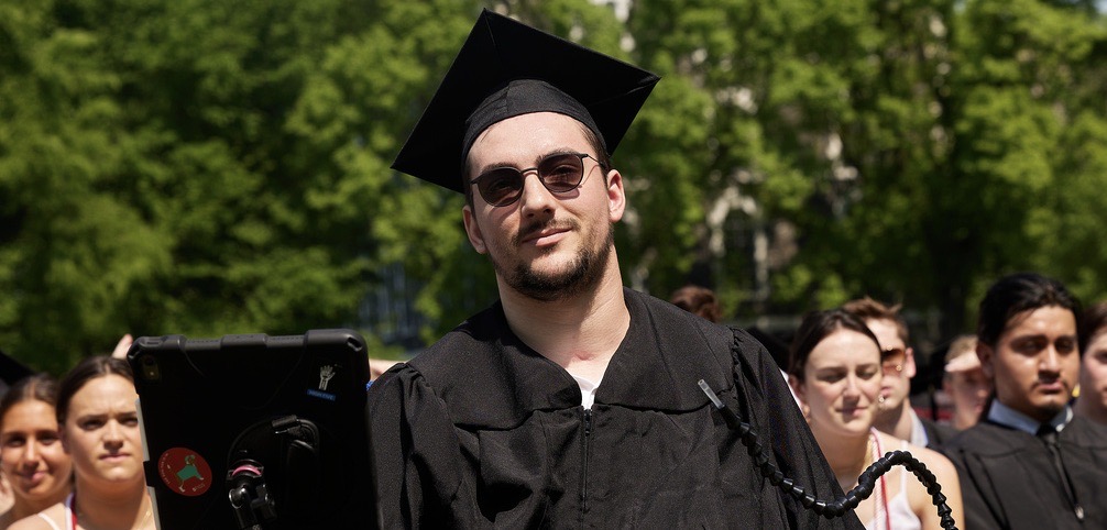 A man wearing sunglasses and a black graduation cap and gown smiles.