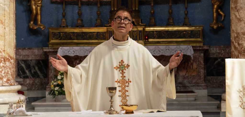 A man holds out his hands, while wearing a white priestly gown.