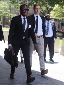 A man wearing a business suit and holding a suitcase strides on a street while smiling.