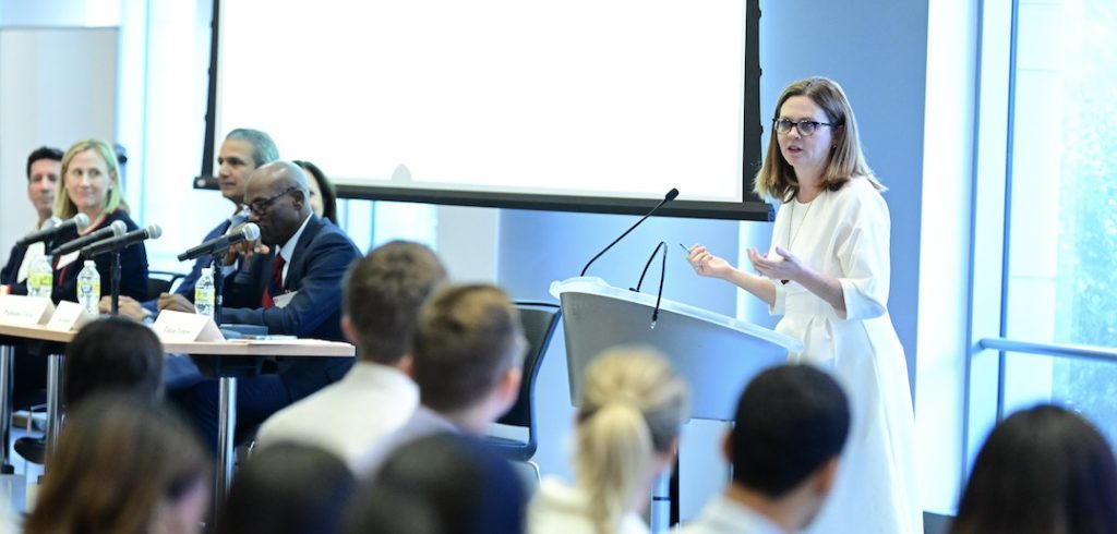 A woman speaks in front of a podium while seated people in the background look on.